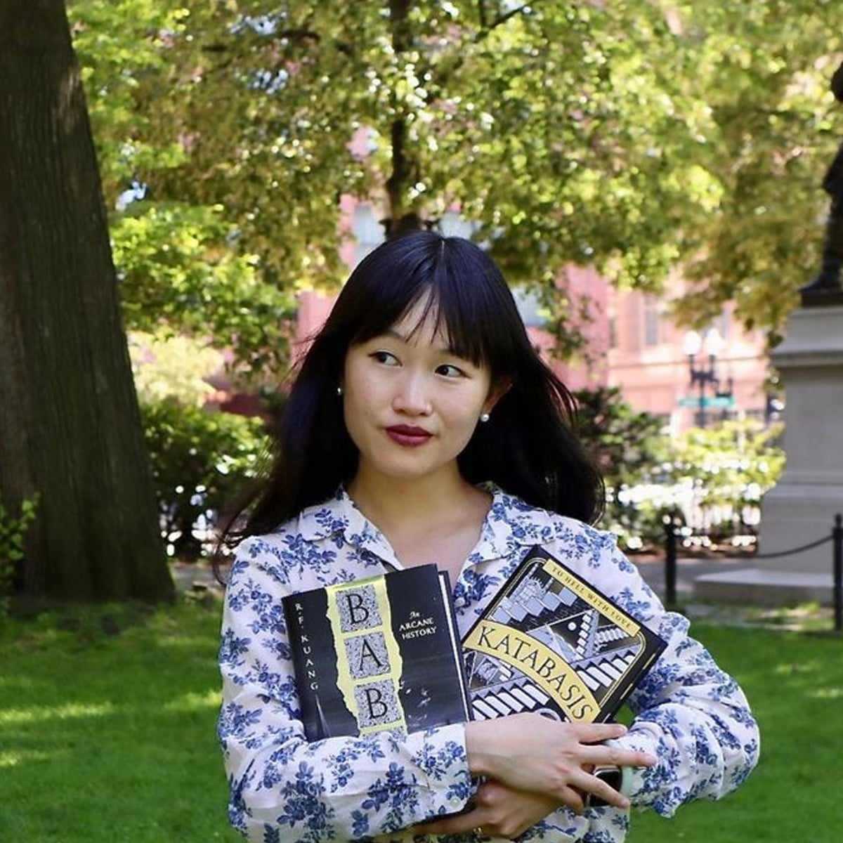A woman with long dark hair and a floral dress stands outdoors, holding two books titled "BAB" and "KATABASIS," with trees and sunlight in the background.