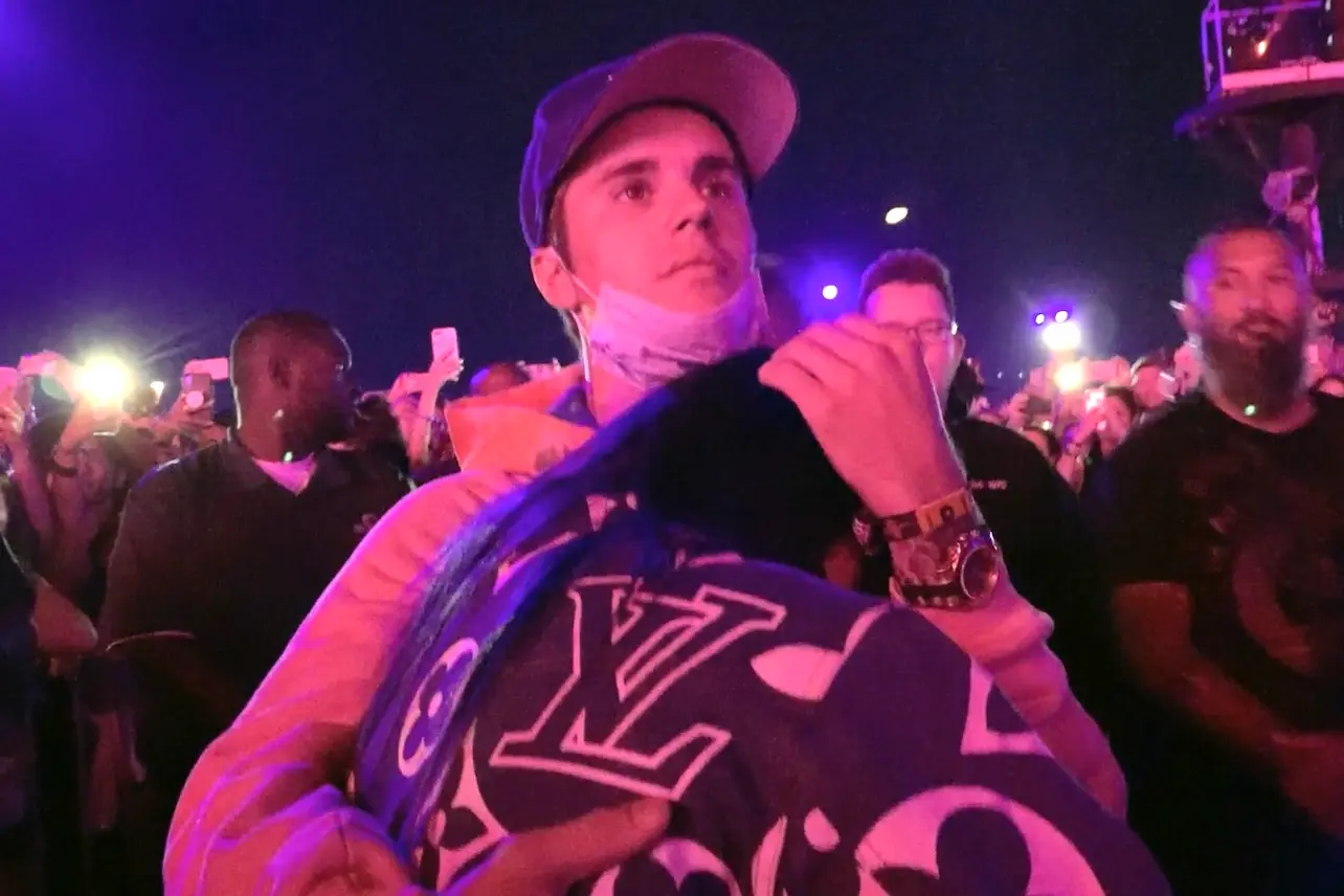 A young man wearing a cap and a mask under his chin holds a person wrapped in a blue Louis Vuitton blanket in a crowd at night, surrounded by people and phone lights.