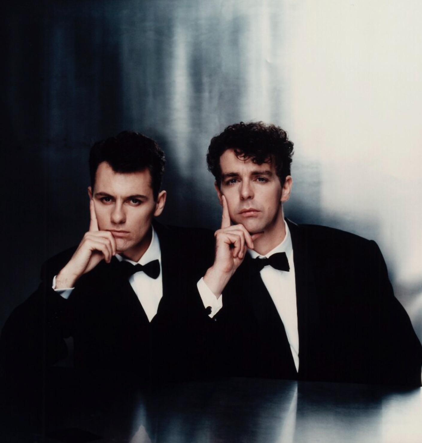 Two men in black tuxedos sit side by side against a metallic background, each resting their chin on one hand in a thoughtful pose, looking directly at the camera with serious expressions.