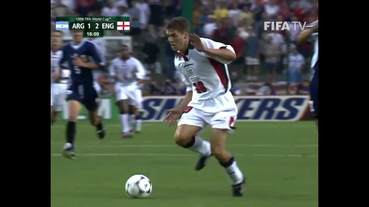A soccer player in a white and red England kit runs with the ball during a match against Argentina, with the scoreboard showing ARG 1-2 ENG. The field and crowd are visible in the background.