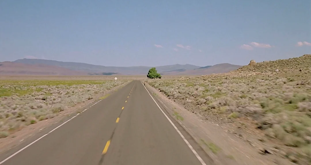 A straight, empty two-lane road stretches into the distance through a dry, open desert landscape under a clear blue sky, with low mountains visible on the horizon.