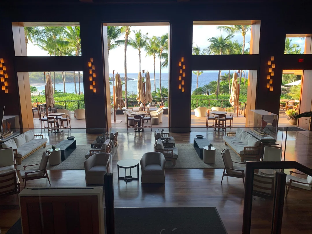 Spacious, elegant hotel lounge with modern seating, large windows revealing palm trees, umbrellas, and a scenic ocean view under a sunny sky.