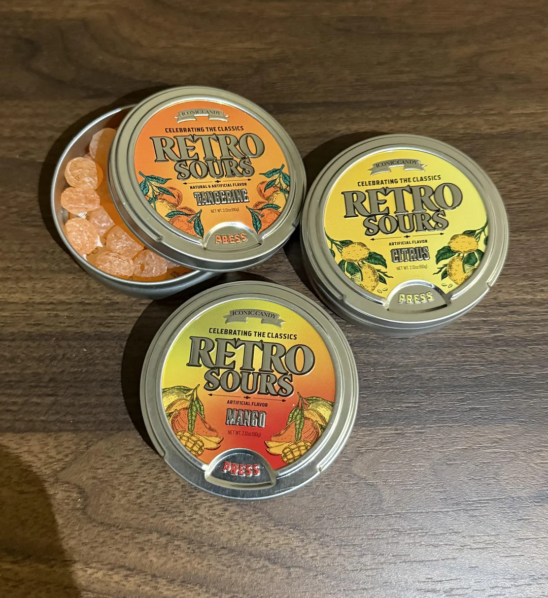 Three round tins of “Retro Sours” candy, with two closed (Mango and Lychee flavors) and one open (showing orange candies inside) are on a dark wooden surface. The tins have vintage-style labels.