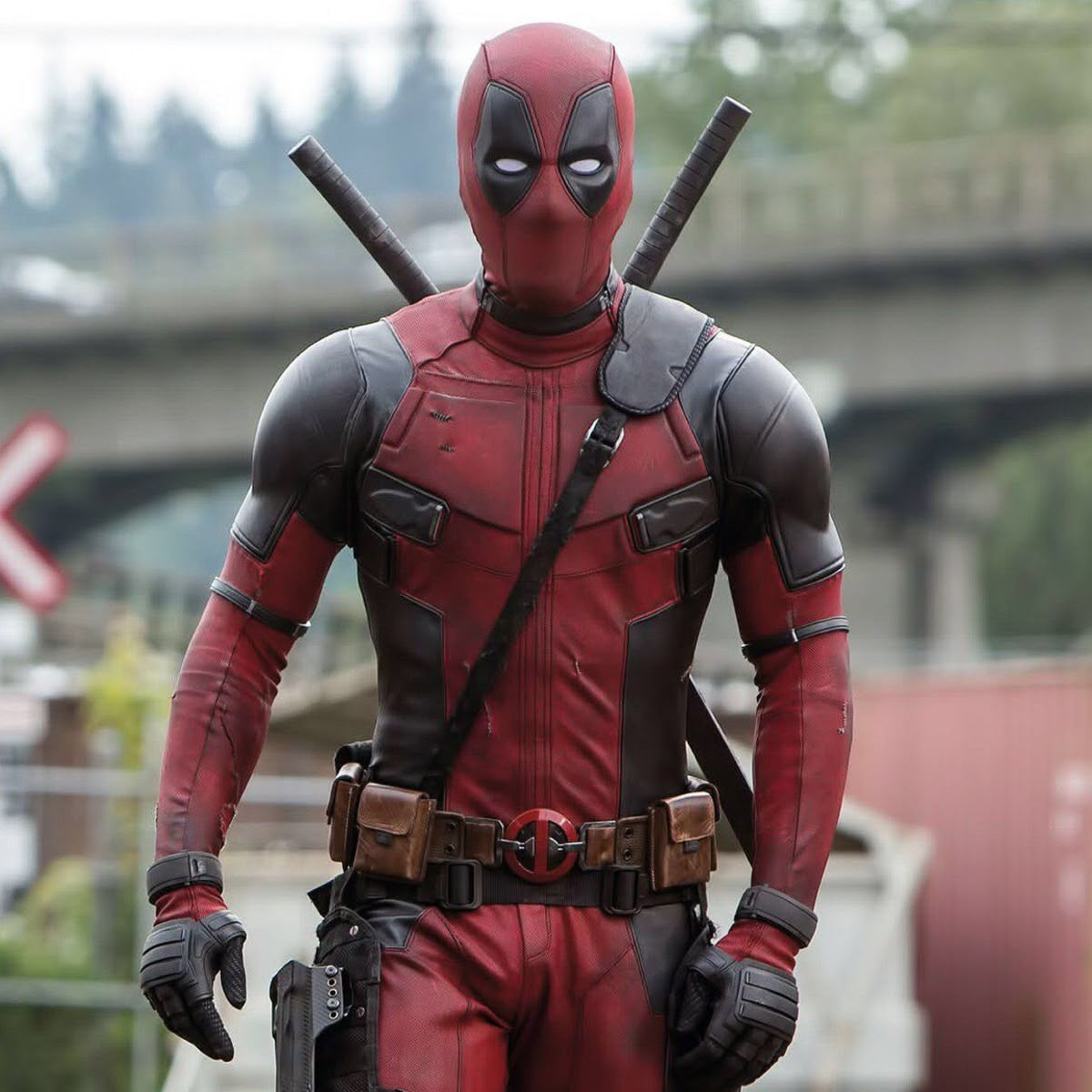 A person in a red and black superhero costume with two swords on their back stands outdoors, with a blurred bridge and trees in the background.