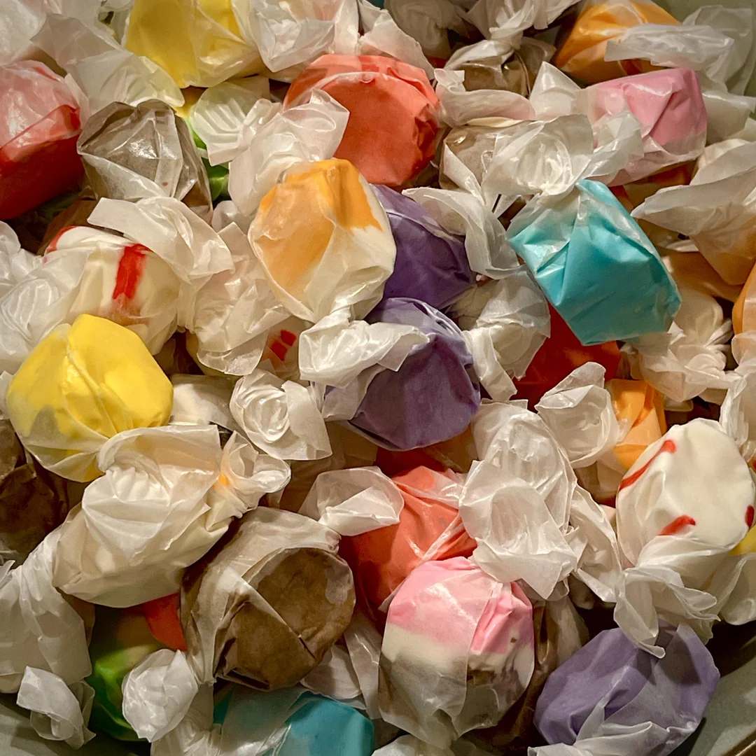 Assorted pieces of saltwater taffy in colorful wrappers, including yellow, blue, purple, orange, red, and brown, are piled together with white twisted ends visible on each piece.