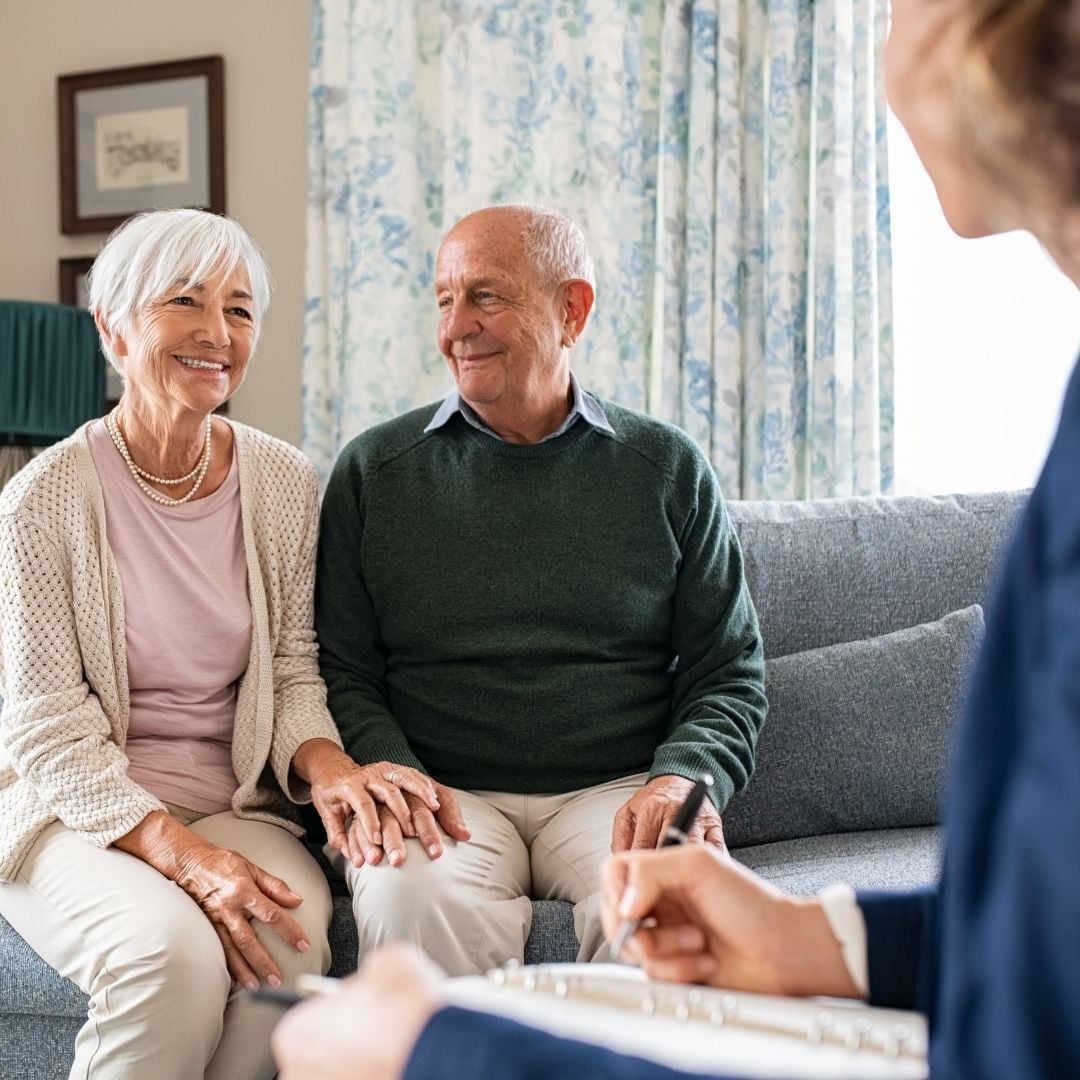 An elderly couple sits closely together on a sofa, smiling and holding hands, while a person in the foreground appears to speak with them, possibly during a consultation or meeting at home.
