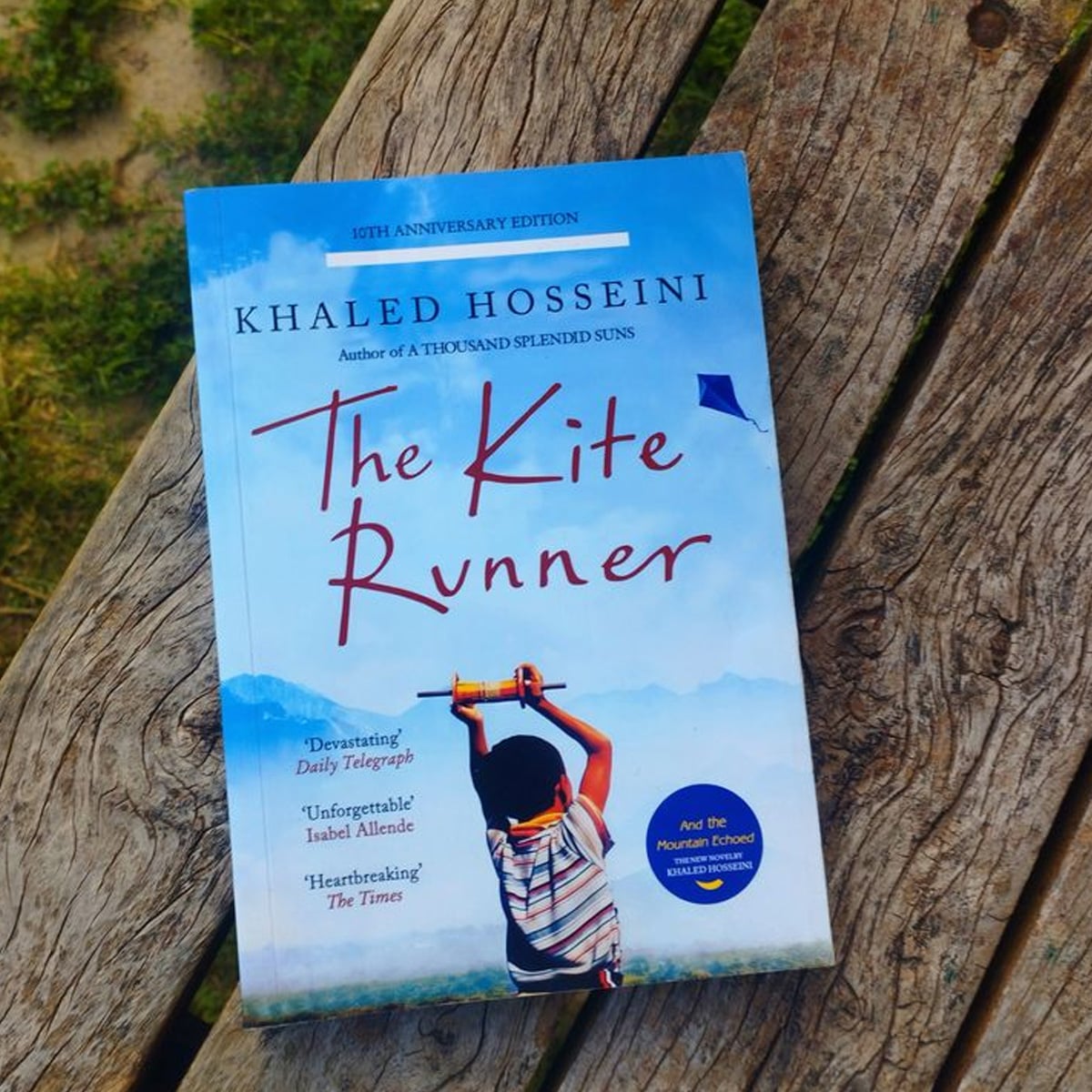A copy of the book "The Kite Runner" by Khaled Hosseini rests on a weathered wooden surface outdoors. The cover features a boy flying a kite against a blue sky.