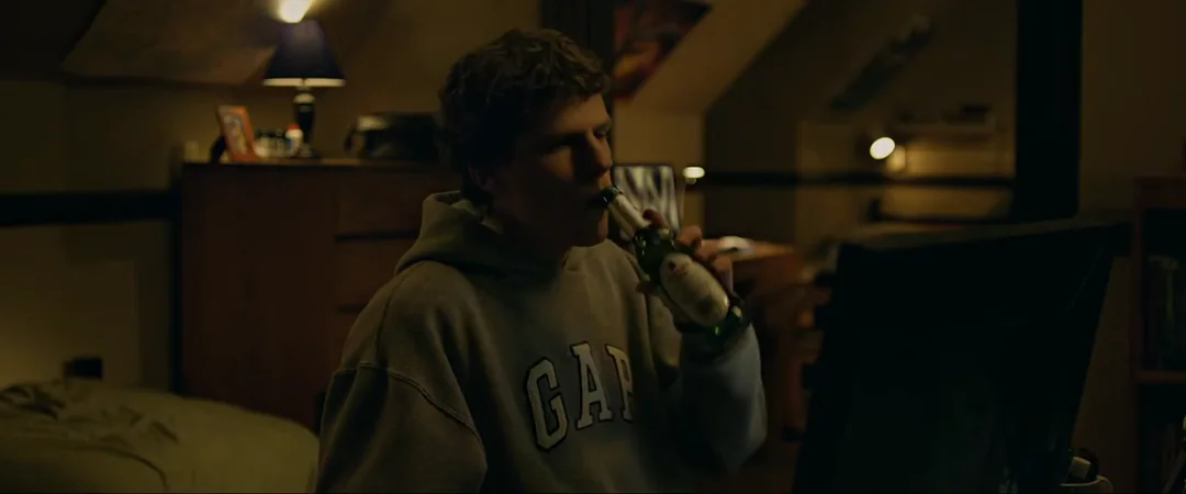 A young man in a gray GAP hoodie drinks from a beer bottle while sitting at a desk in a dimly lit bedroom with a lamp and bed in the background.