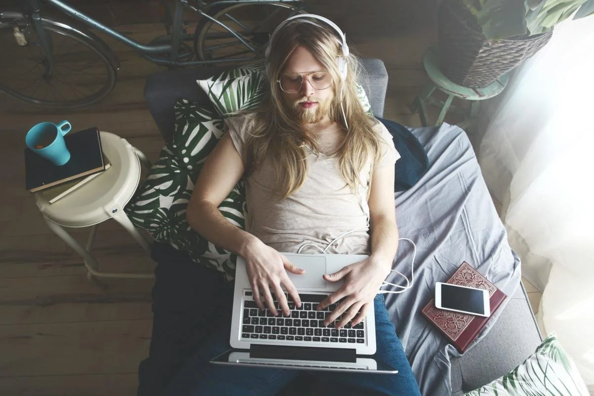 A person with long hair, wearing glasses and headphones, lies on a sofa using a laptop. Nearby are a book, a tablet, a mug, and a bicycle in the background, with sunlight streaming through a window.
