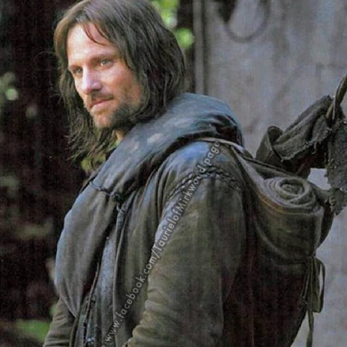 A man with long brown hair and a beard stands outdoors wearing rugged, dark clothing and a rolled-up blanket or cloak on his back, looking off to the side with a serious expression.