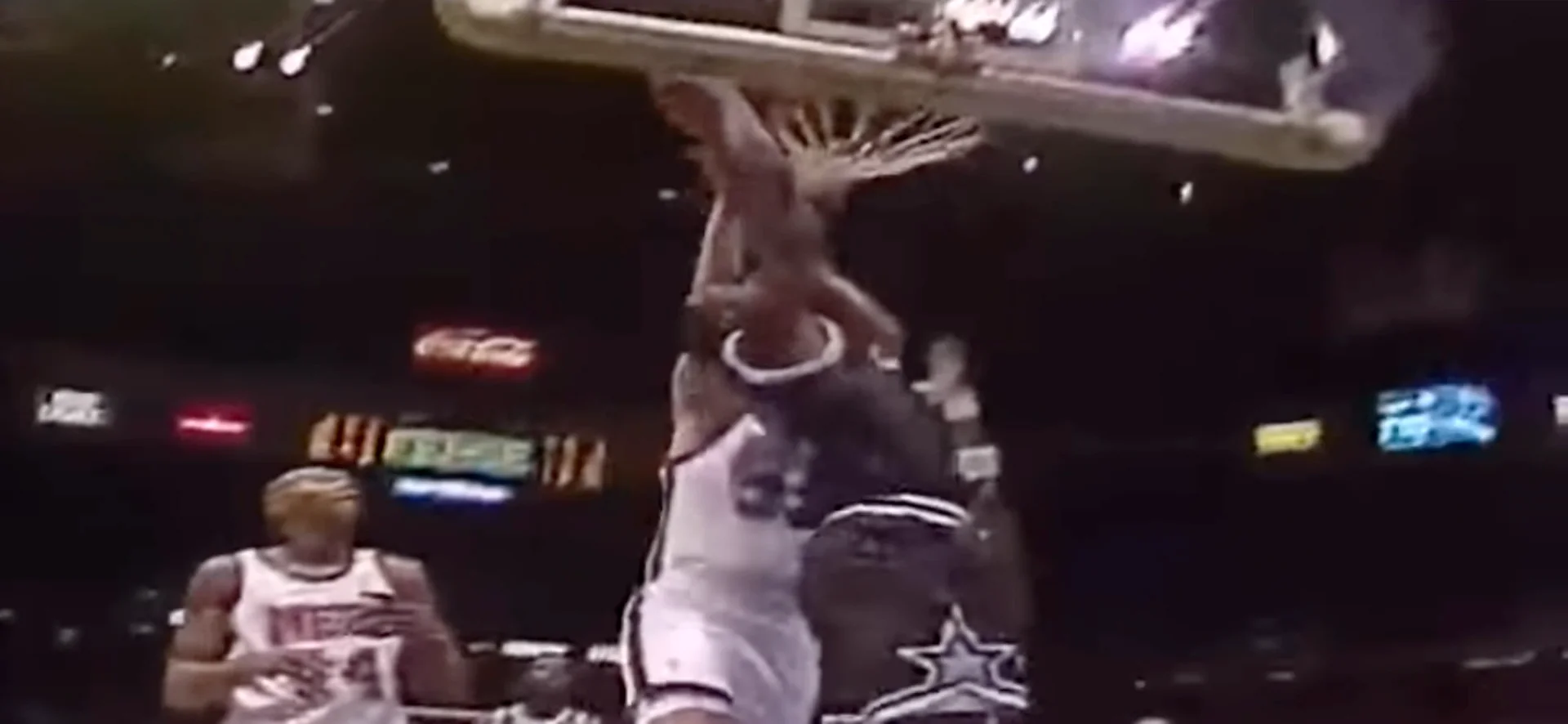 A basketball player in a white uniform powerfully dunks the ball over a defender in a dark uniform, while another player watches nearby. The action takes place under a brightly lit hoop during a game.