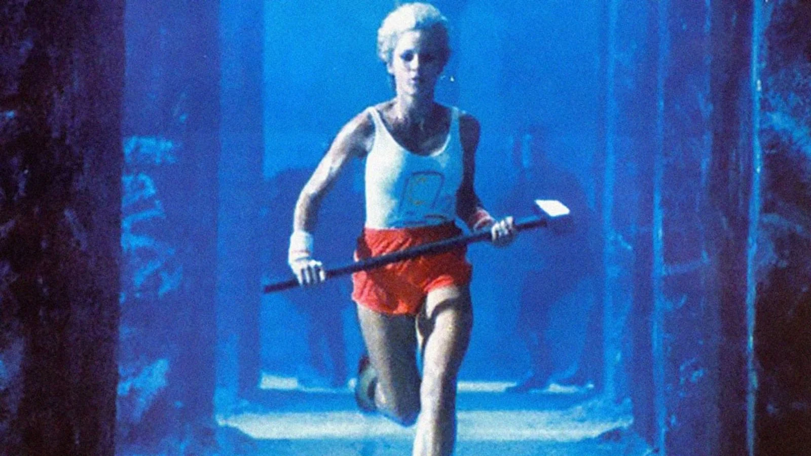 A woman with short blonde hair runs through a blue-lit corridor holding a large hammer. She wears a white tank top, red shorts, and white athletic shoes. The background is dark and shadowy.