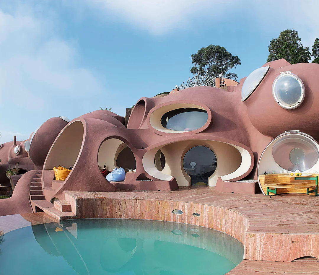 A futuristic house with rounded, dome-like shapes and large circular windows, set against a blue sky. There is a curved swimming pool and wooden deck with outdoor seating in front of the unique pinkish-brown structure.