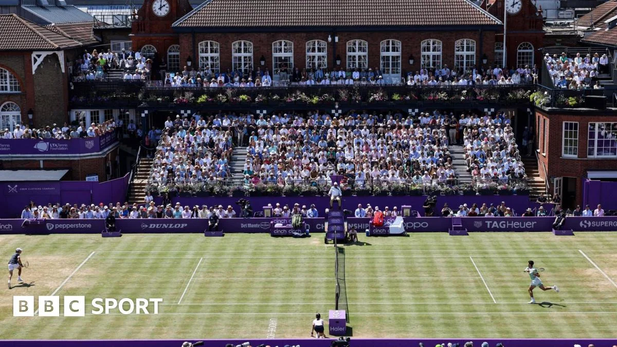 A grass tennis court during a match, with two players competing. The stands are filled with spectators and a historic brick clubhouse is in the background. BBC Sport branding appears in the lower left corner.