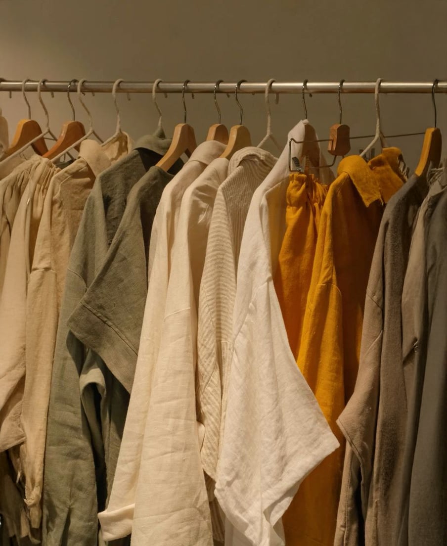 A clothing rack displays various loose, linen garments in neutral and earthy tones, including beige, off-white, olive green, and mustard yellow, all hanging neatly on wooden hangers.