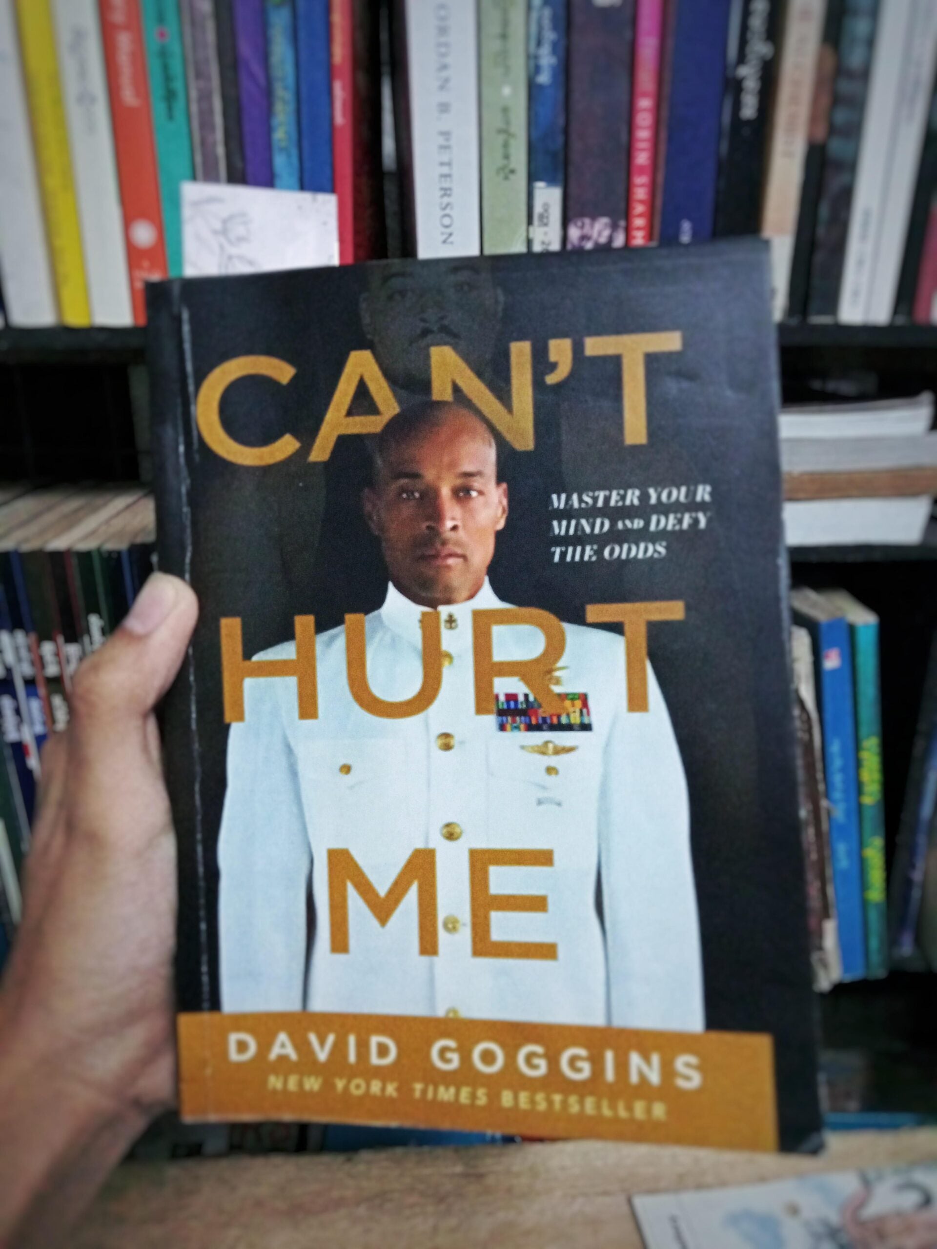 A hand holds the book "Can't Hurt Me" by David Goggins, featuring a portrait of Goggins in a white military uniform. Bookshelves with various colorful books are visible in the background.