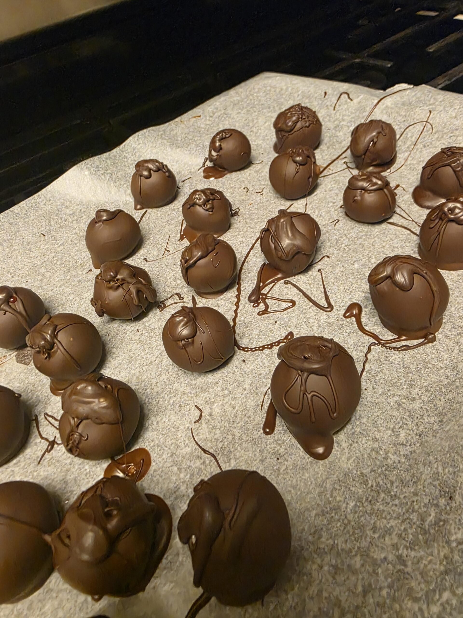 Rows of round chocolate truffles are arranged on a baking sheet lined with parchment paper, with chocolate drizzled over some of the truffles.
