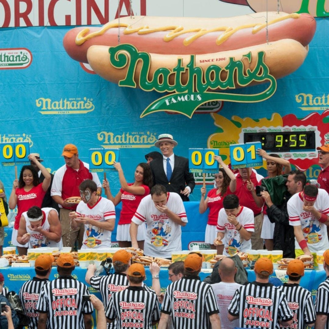 A group of people participate in Nathan's Famous hot dog eating contest, with judges holding scorecards, a large hot dog sign overhead, and referees in striped shirts watching the competitors.