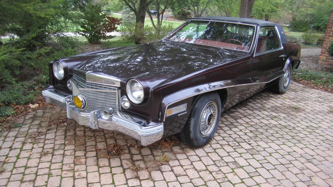 A vintage maroon 1970s Cadillac Eldorado parked on a cobblestone driveway, surrounded by greenery and trees.