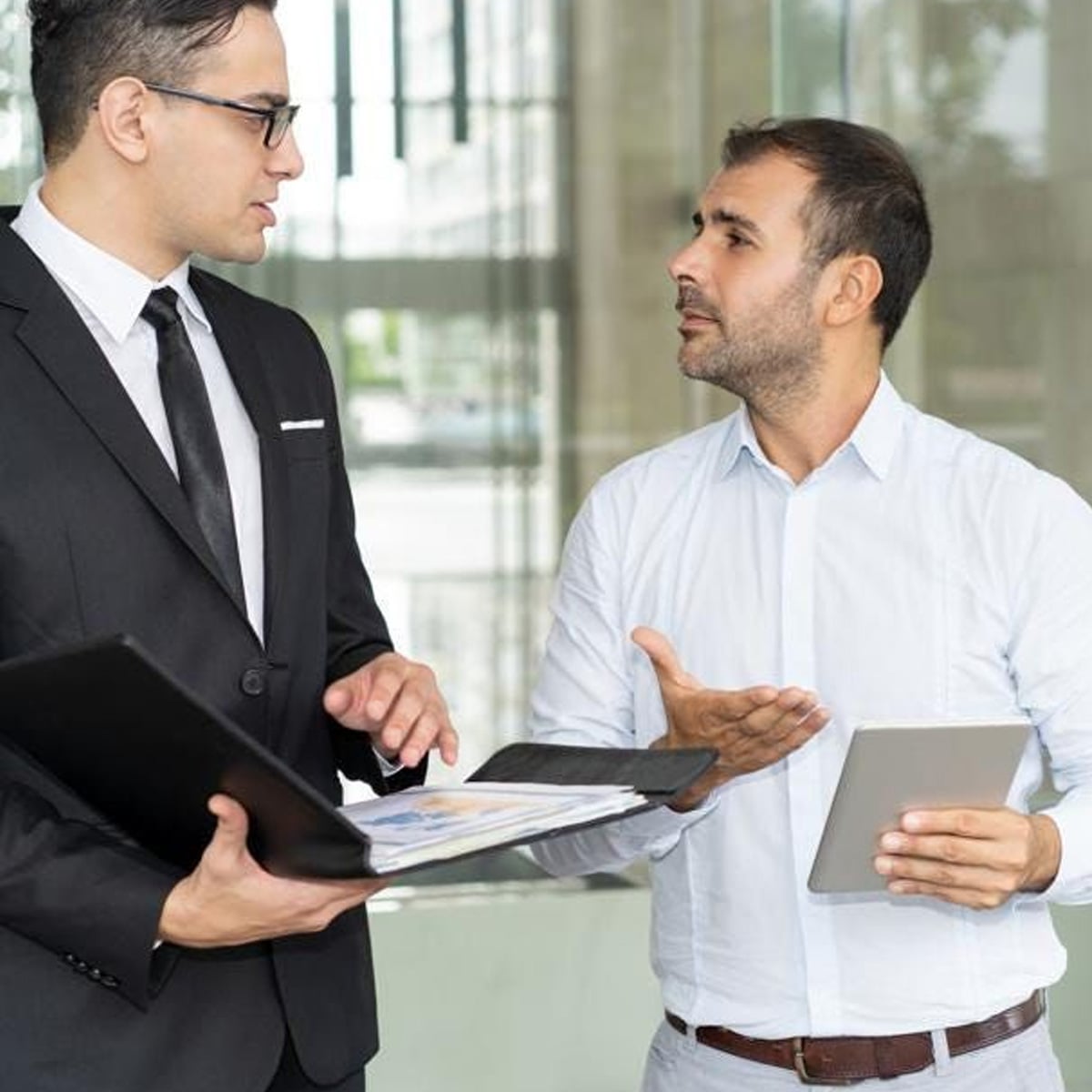 Two men standing indoors having a discussion; one is in a suit holding a folder with documents, the other in a white shirt holding a tablet and gesturing with his hand.