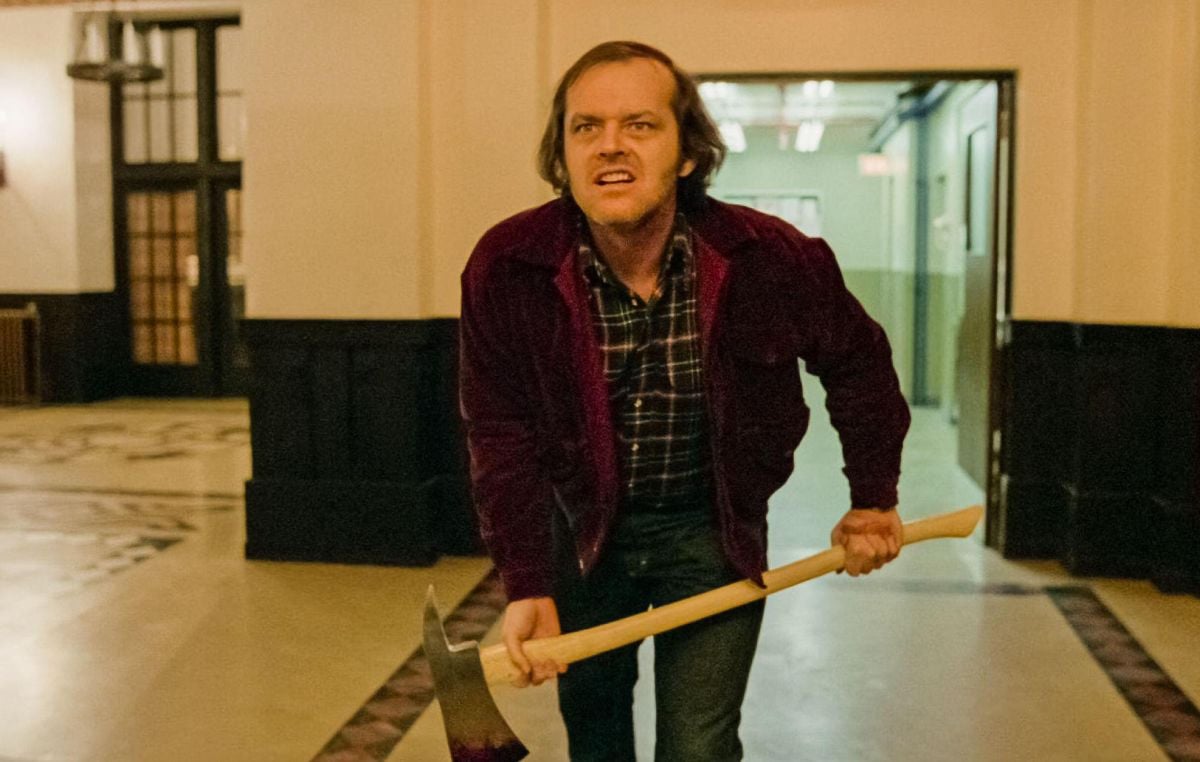 A man with a wild expression, wearing a red jacket and plaid shirt, crouches slightly while holding an axe in both hands inside a large, dimly lit hallway.