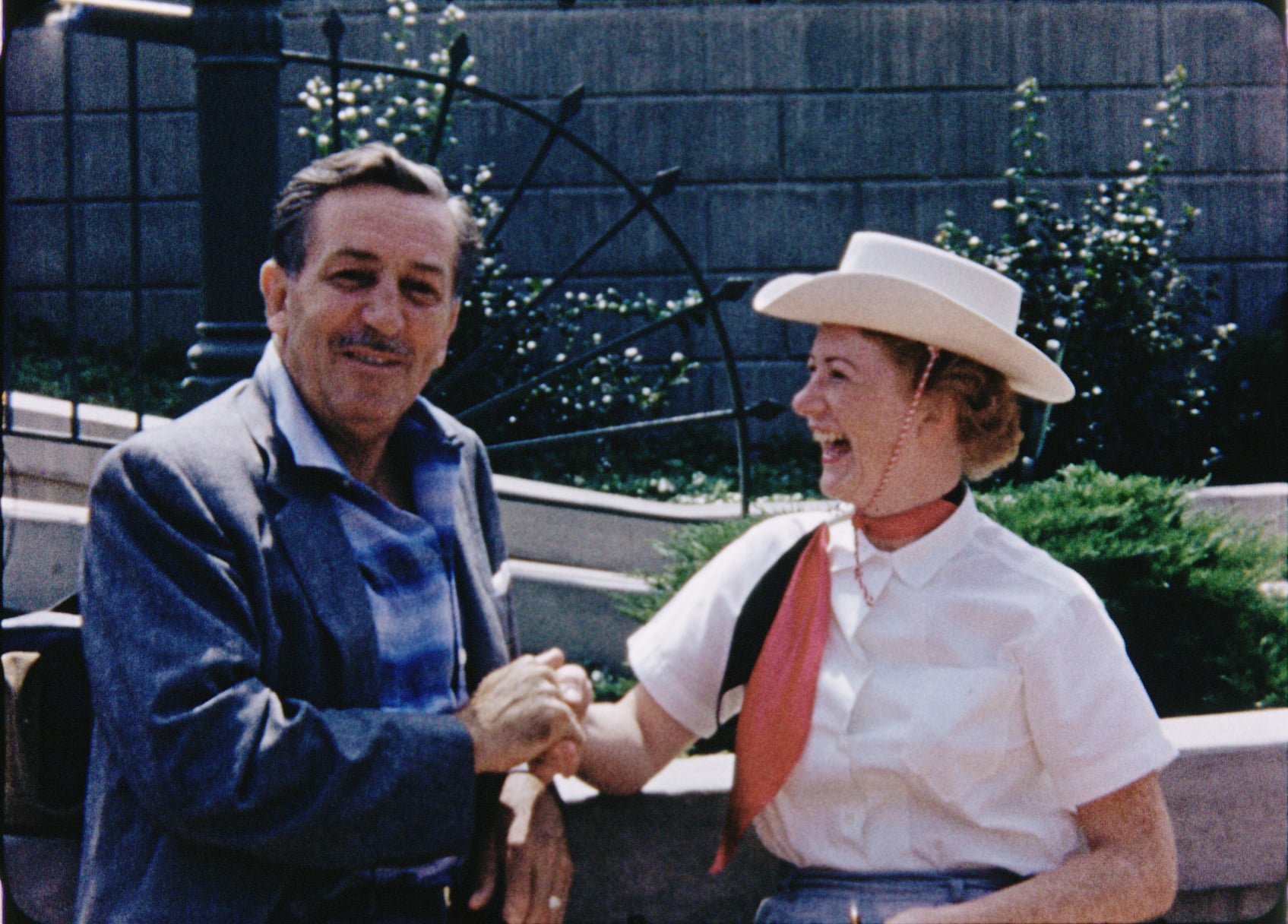 A man in a gray jacket smiles while shaking hands with a woman in a cowboy hat and pink scarf. They are outdoors, standing in front of a decorative fence and greenery.