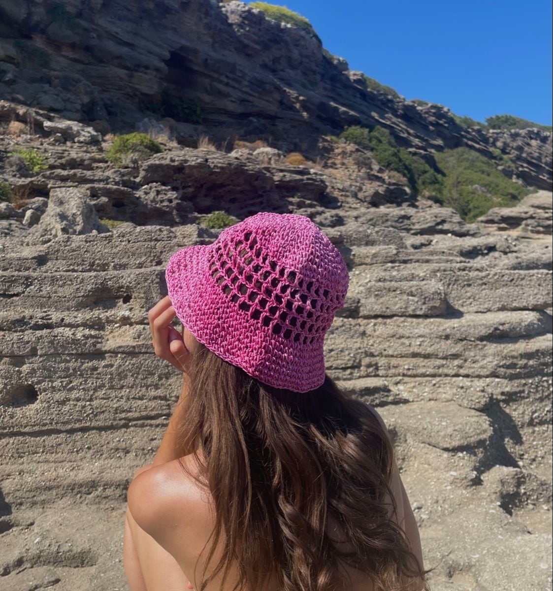 A woman with long brown hair, wearing a pink crochet hat, sits facing rocky terrain under a clear blue sky. The photo is taken from behind, showing her back and the textured rocks in the background.