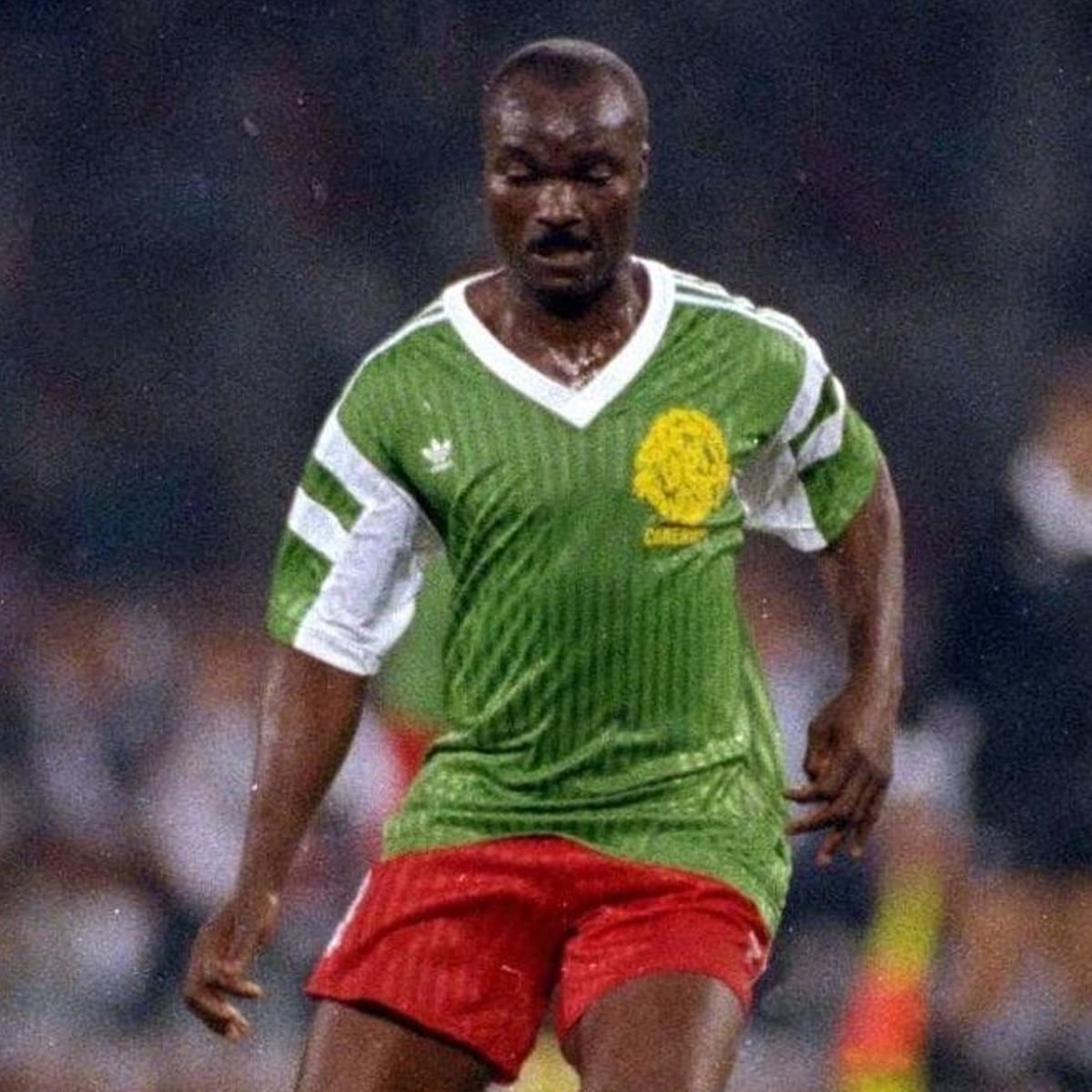 A football player in a green and white Cameroon jersey with red shorts is running on the field during a match. The background is blurred, emphasizing the player’s movement and focus.