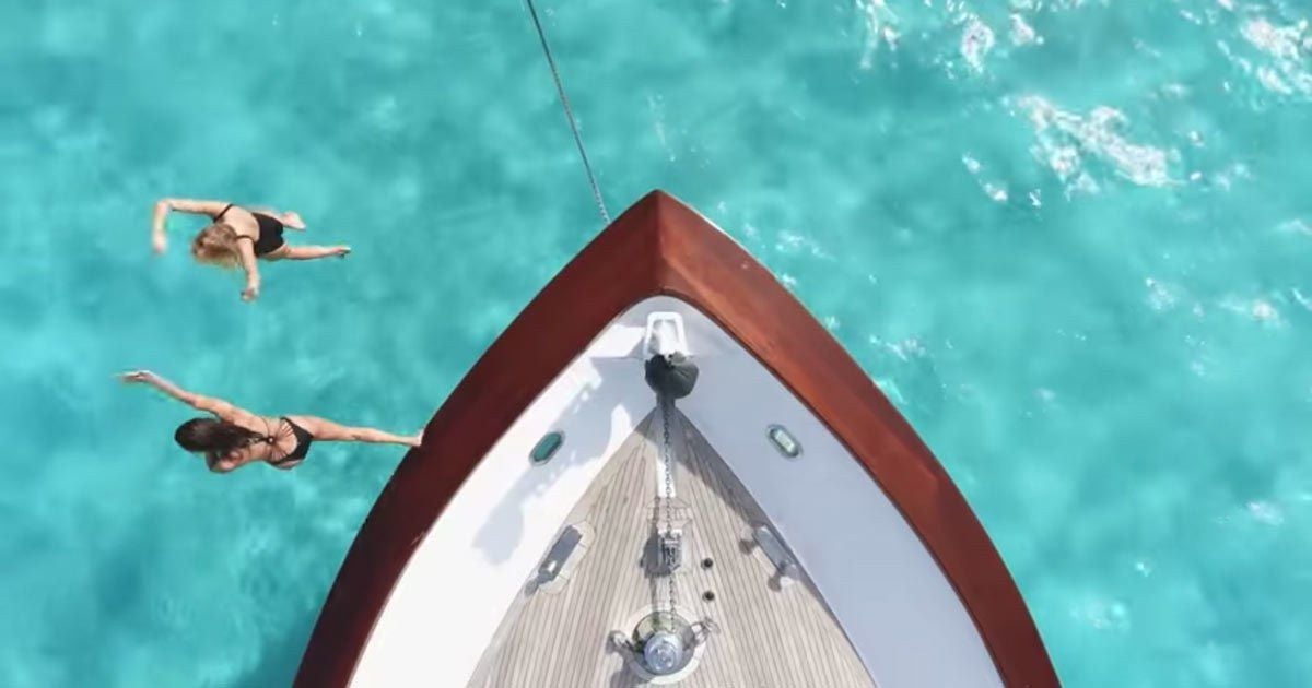 Aerial view of the bow of a yacht anchored in clear turquoise water, with two women in swimsuits swimming nearby.
