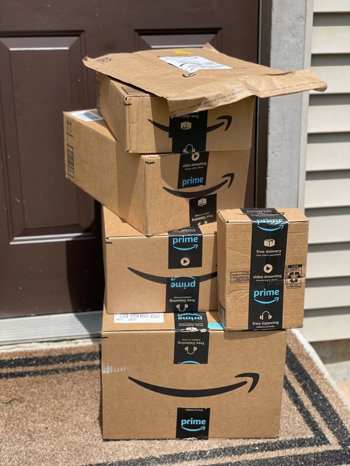 A stack of five Amazon Prime packages, including one damaged box, is placed on a doormat in front of a brown door and beige siding.