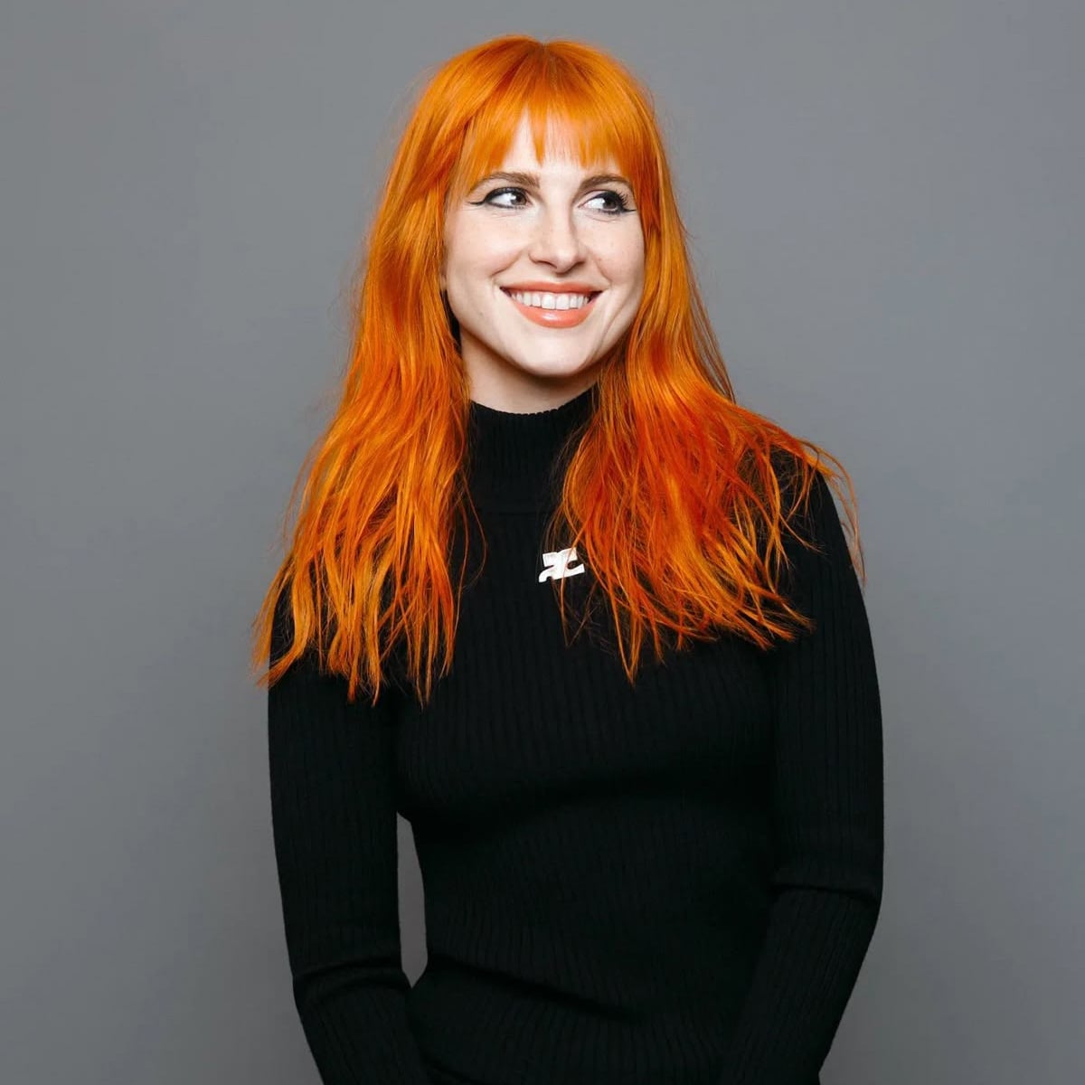 A person with bright orange hair and bangs, wearing a long-sleeved black ribbed top, smiles while looking slightly to the side against a plain gray background.