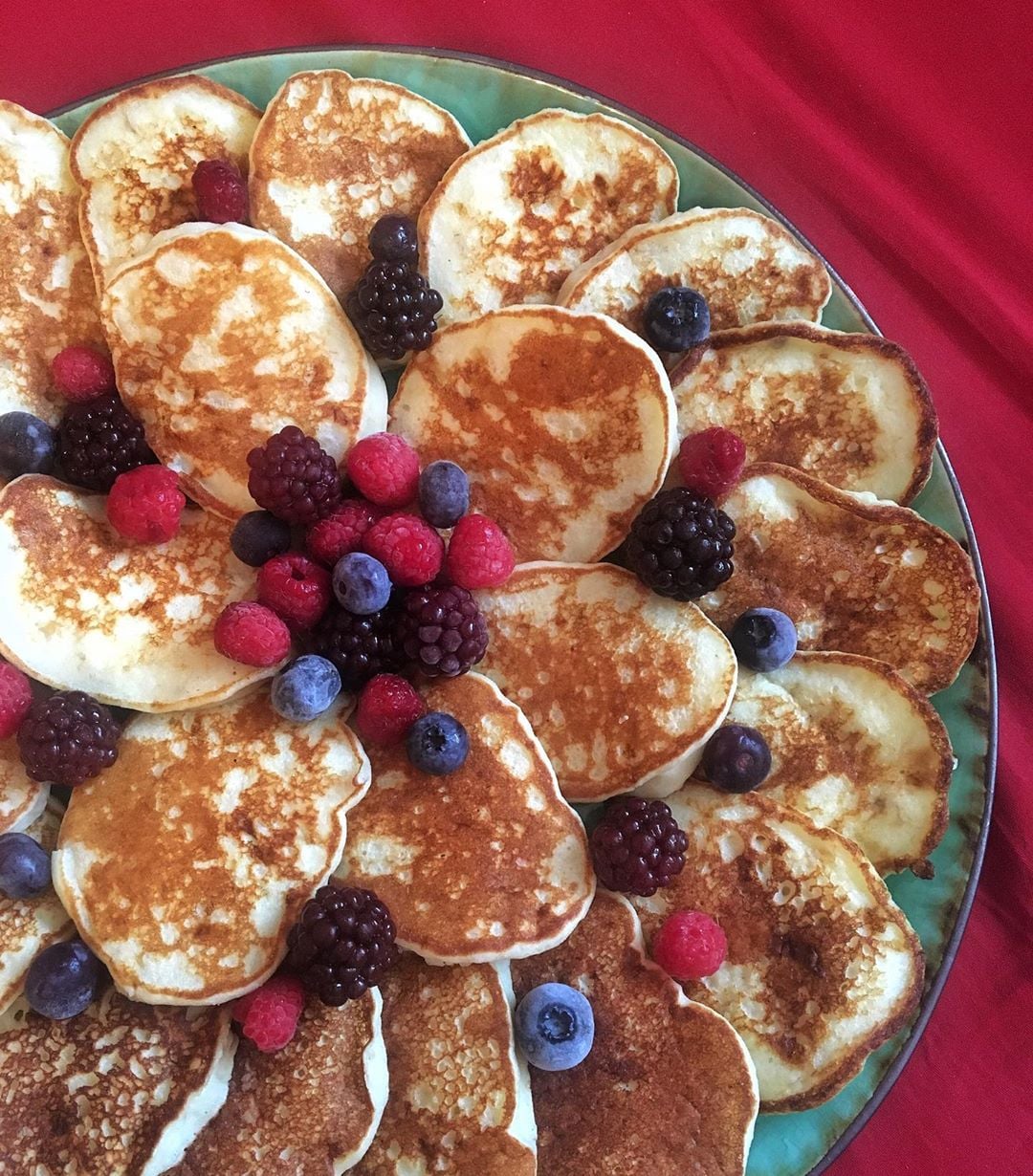 A large plate of golden pancakes arranged in a circular pattern, topped with clusters of mixed berries including blackberries, raspberries, and blueberries, on a red tablecloth.