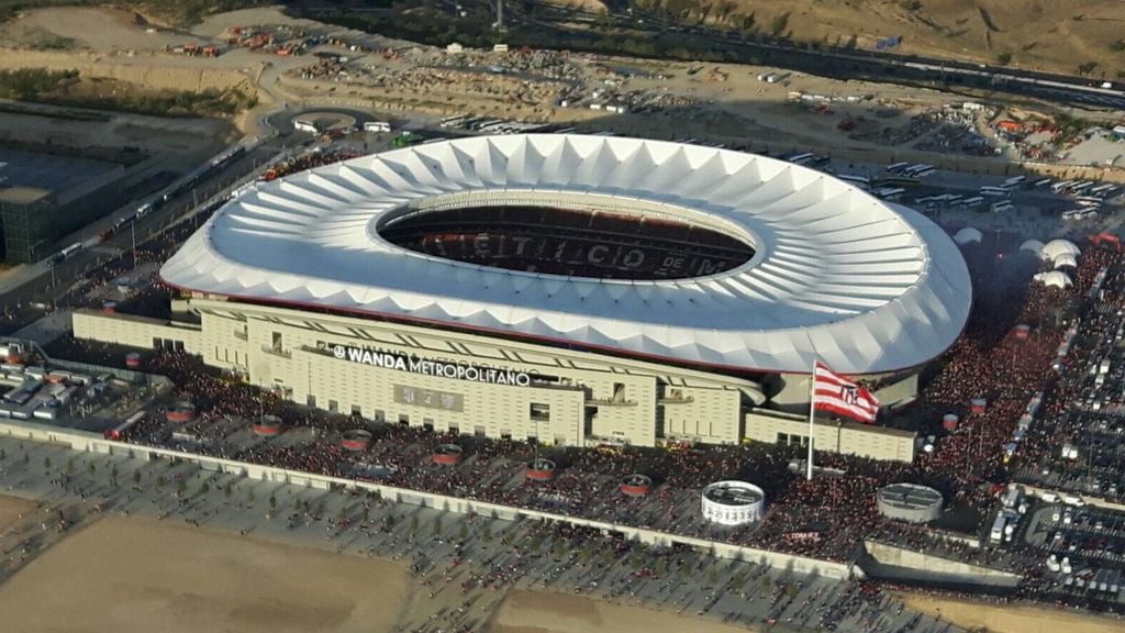 Aerial view of a large, modern stadium packed with people. The stadium has a unique white, oval-shaped roof and the name "Wanda Metropolitano" is visible on the facade. A large flag stands in front.