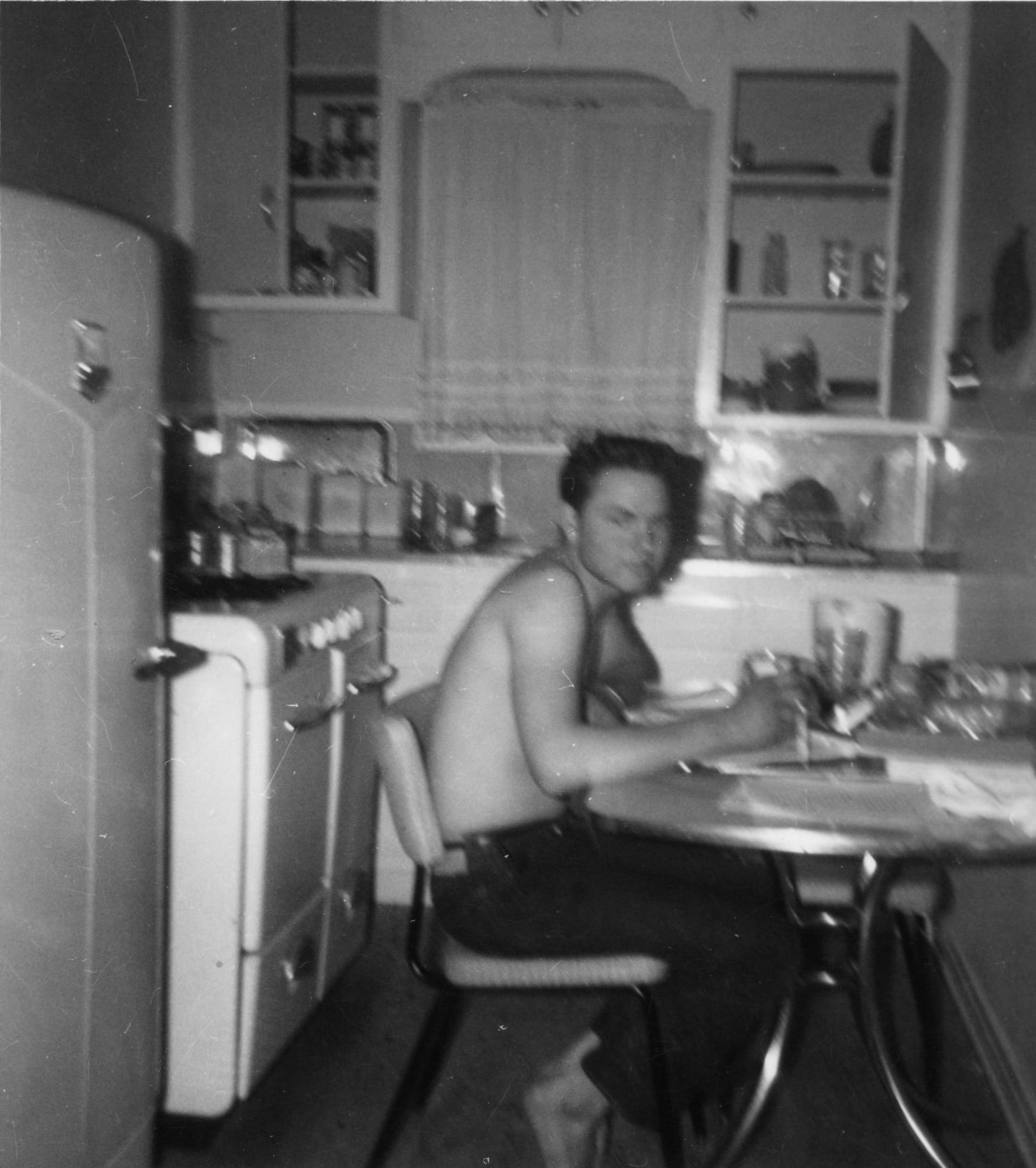 A black-and-white photo of a shirtless person sitting at a kitchen table, mid-meal. The vintage kitchen has an old refrigerator, stove, cabinets, and shelves with various items. The person looks toward the camera.