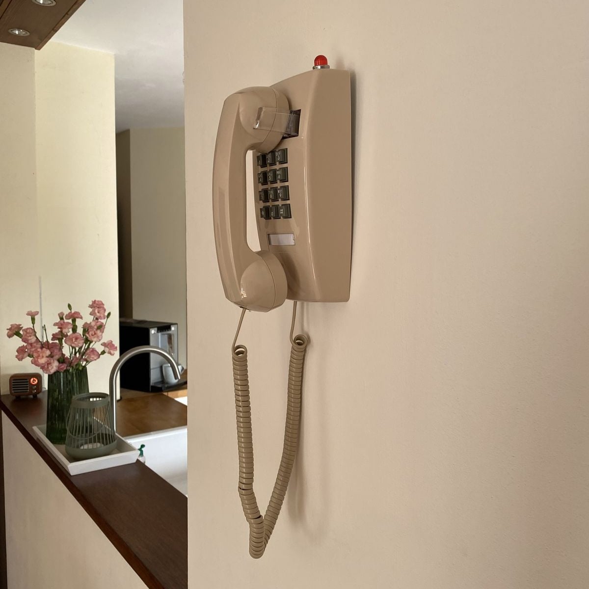 A beige wall-mounted rotary-style telephone with a coiled cord is attached to a light-colored wall. In the background, there are pink flowers in a vase and a modern kitchen sink.