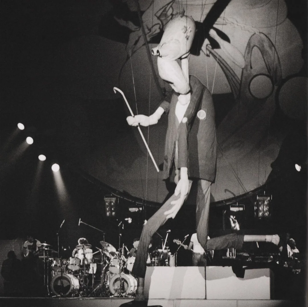 A large, exaggerated puppet figure in a suit and holding a cane dominates the stage, with a rock band performing in the background under dramatic lighting.