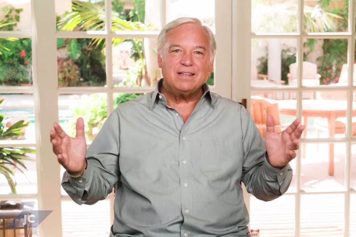 A man with gray hair, wearing a light gray button-up shirt, sits indoors with his hands raised, gesturing while speaking. Behind him are large glass doors showing a bright, leafy outdoor patio.
