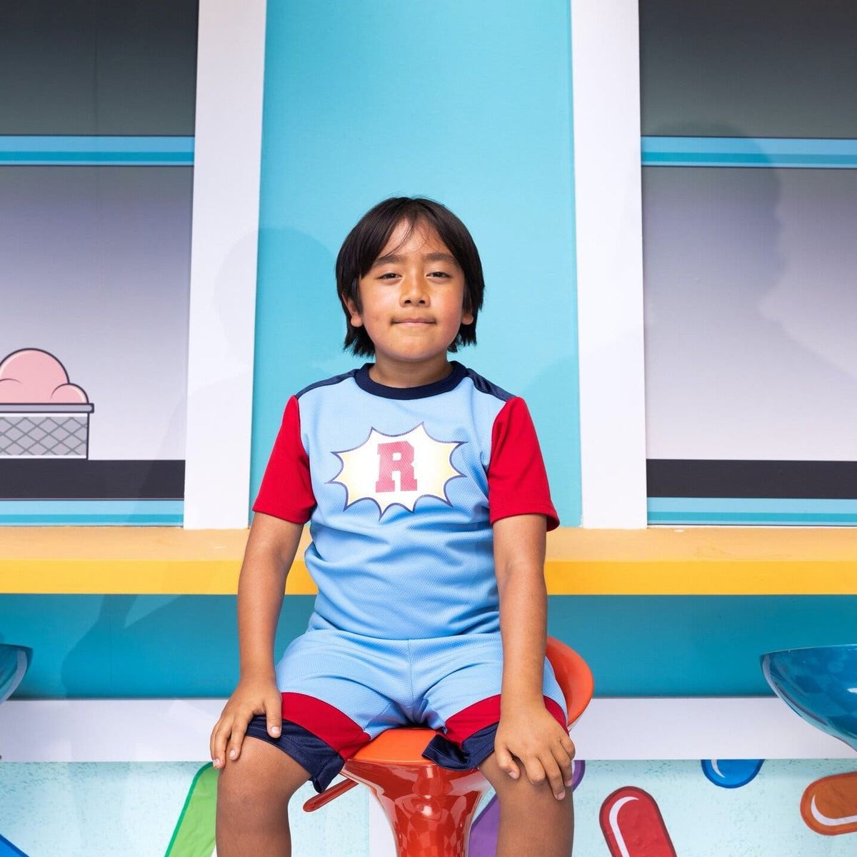 A young boy with medium-length dark hair sits on an orange stool, wearing a blue and red outfit with a white shield and red "R" on the front. He is in front of a colorful wall with windows and a drawing of an ice cream cone.