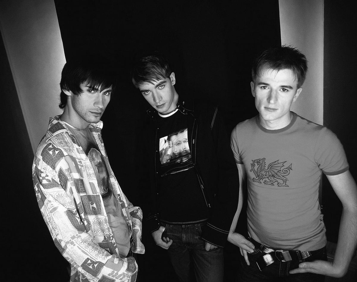 Three young men pose together indoors against a dark backdrop. The image is black and white; the men have serious expressions and are dressed in casual clothes with varied patterns and graphics.