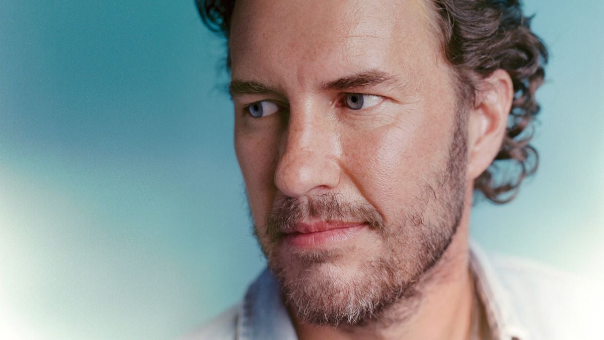 A man with wavy brown hair, a beard, and blue eyes looks thoughtfully to the side against a soft blue background. He is wearing a light-colored shirt.