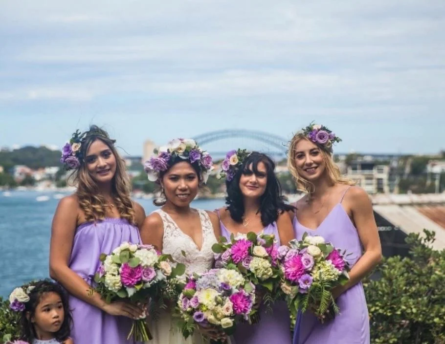 Four women, one in a white dress and three in lavender dresses with floral crowns, hold bouquets of purple and white flowers, standing outdoors by water with a large bridge and cityscape in the background. A young girl is at the side.