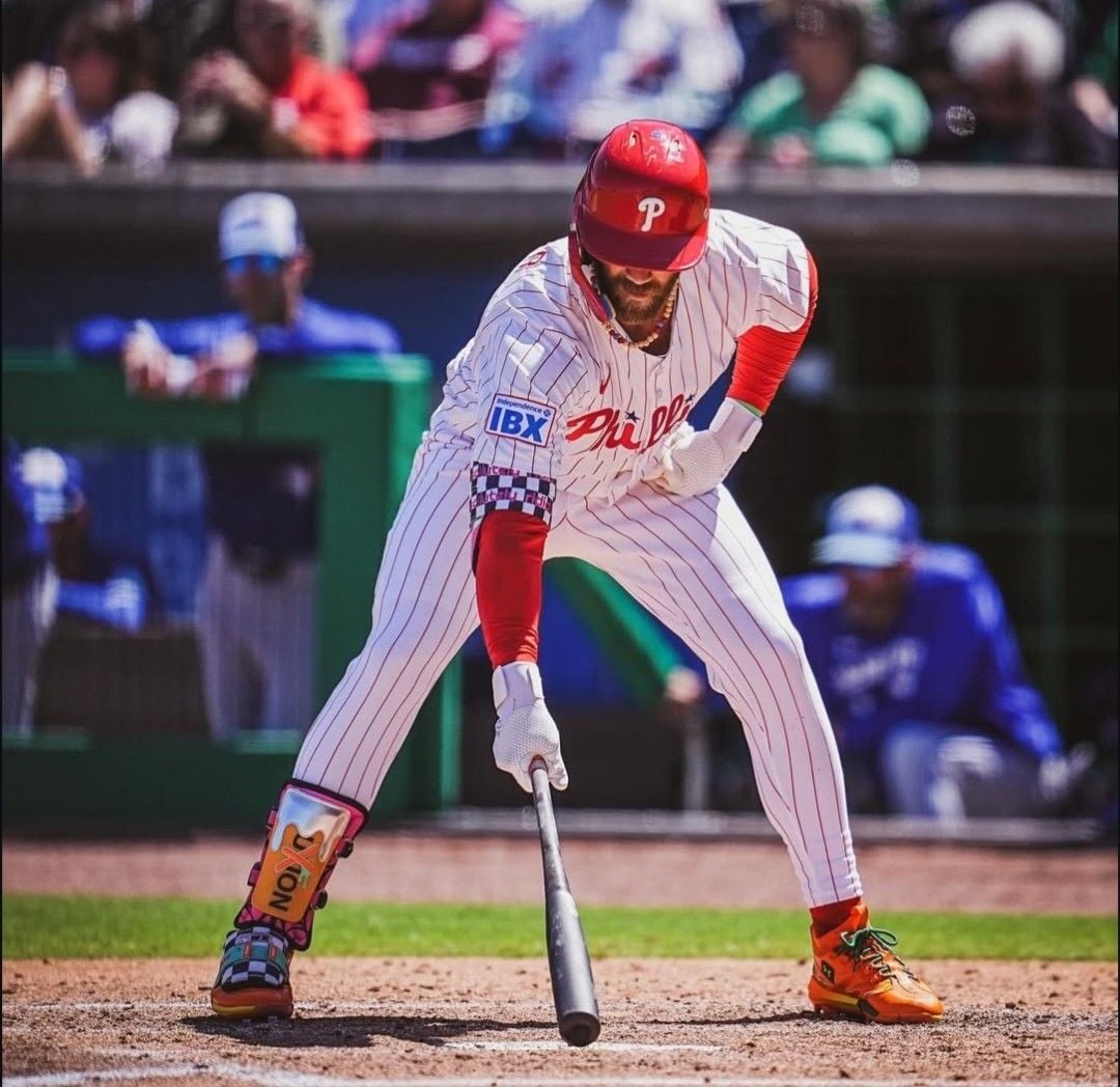 A baseball player in a white and red pinstriped Phillies uniform bunts the ball during a game, with spectators and opposing team members visible in the background.