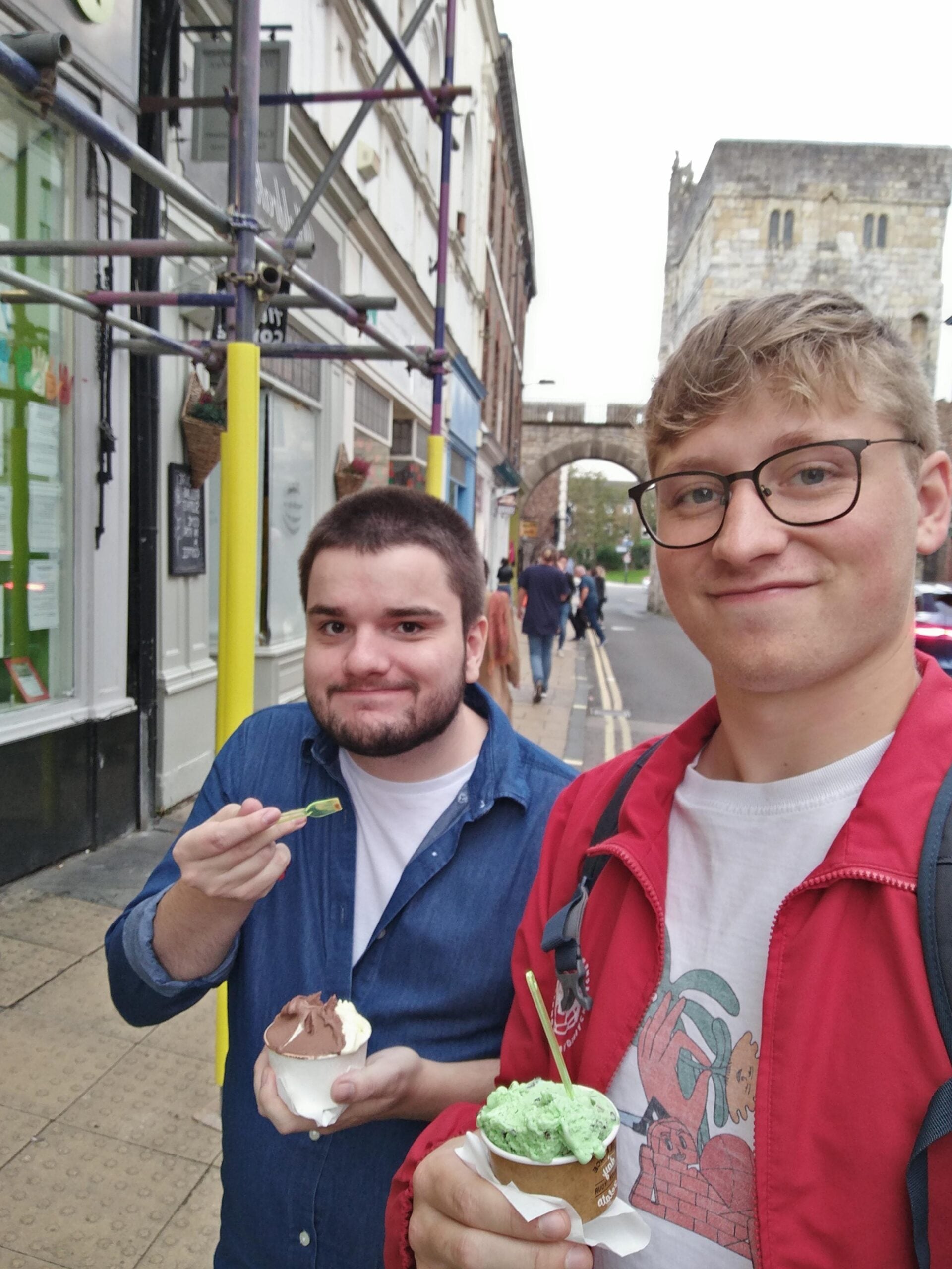 Two men stand on a city sidewalk eating ice cream. One wears glasses and a red jacket holding mint ice cream, while the other wears a blue shirt holding chocolate ice cream. Buildings and an old stone arch are in the background.