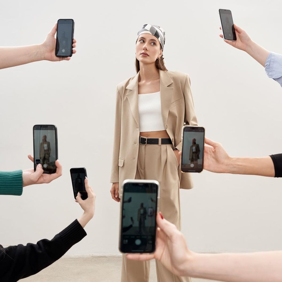 A woman in a beige suit and headscarf poses against a plain background while multiple hands holding smartphones surround her, taking photos or videos.