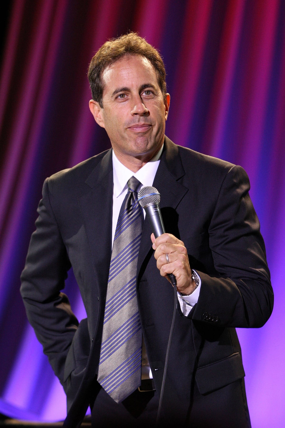 A man in a suit and tie holds a microphone on stage, standing in front of a backdrop with purple and pink lighting.