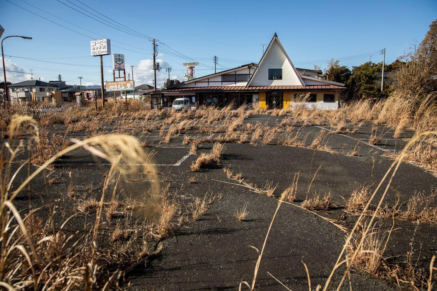 An abandoned restaurant with a triangular roof sits behind an overgrown, cracked parking lot filled with tall, dry grass on a sunny day. Residential houses and utility poles are visible in the background.