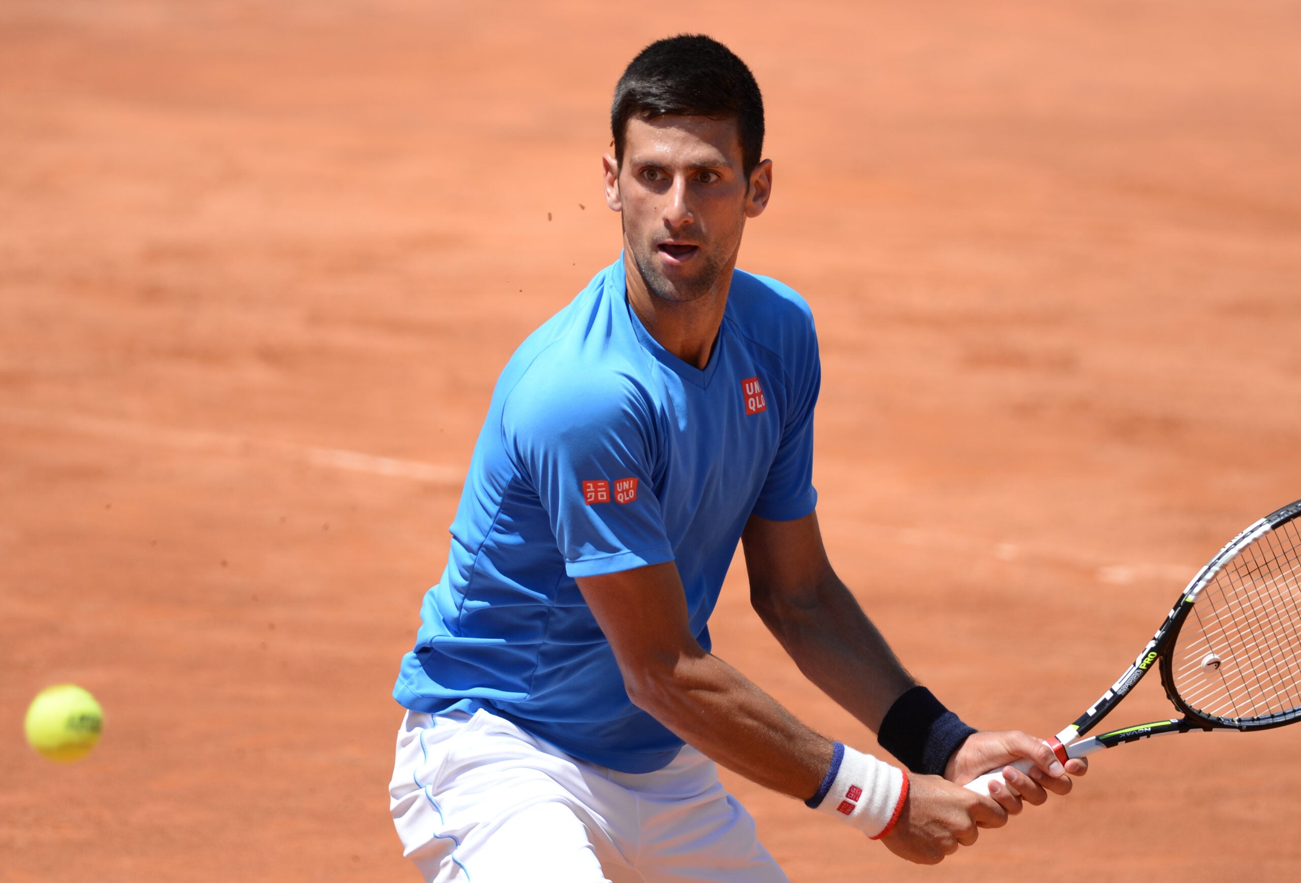 A male tennis player in a blue shirt and white shorts prepares to hit a backhand shot on a clay court, with a tennis ball approaching and a racket in his hands.