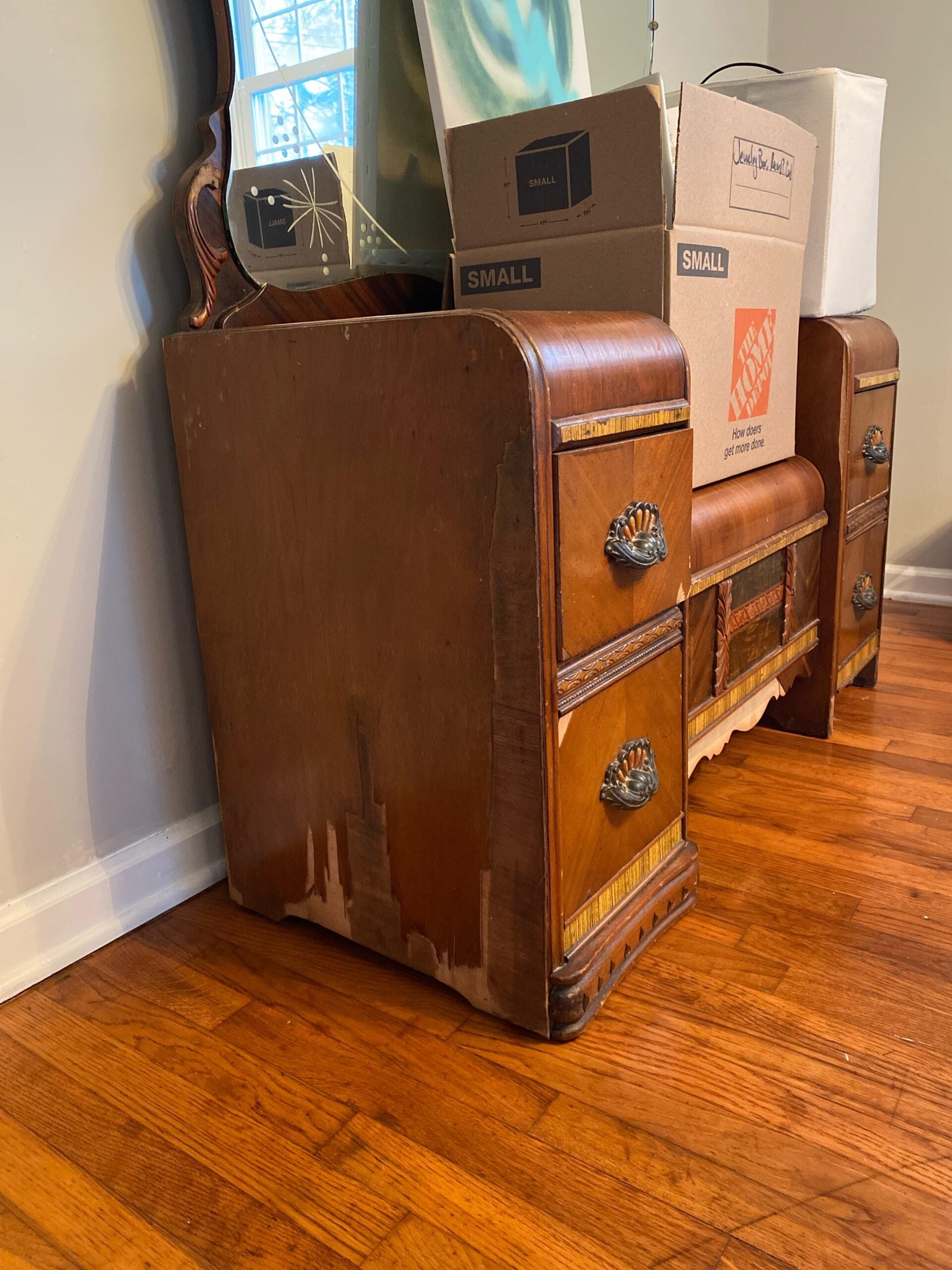 A vintage wooden dresser with some damaged veneer sits on a hardwood floor. There are a mirror, cardboard boxes, and a white container stacked on top of it, against a beige wall.