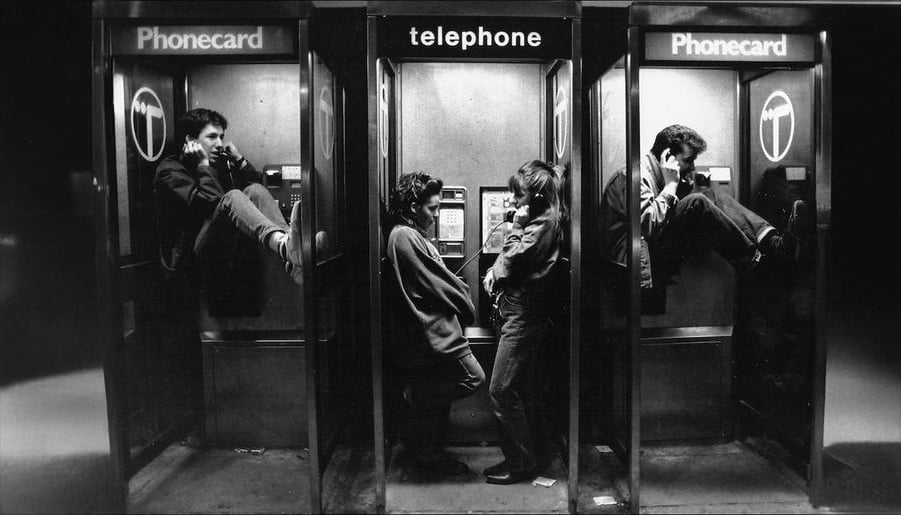 Three phone booths in a row; two people stand and talk in the center booth while two men sit inside the side booths, each using a phone. The scene is in black and white and appears to be from the past.