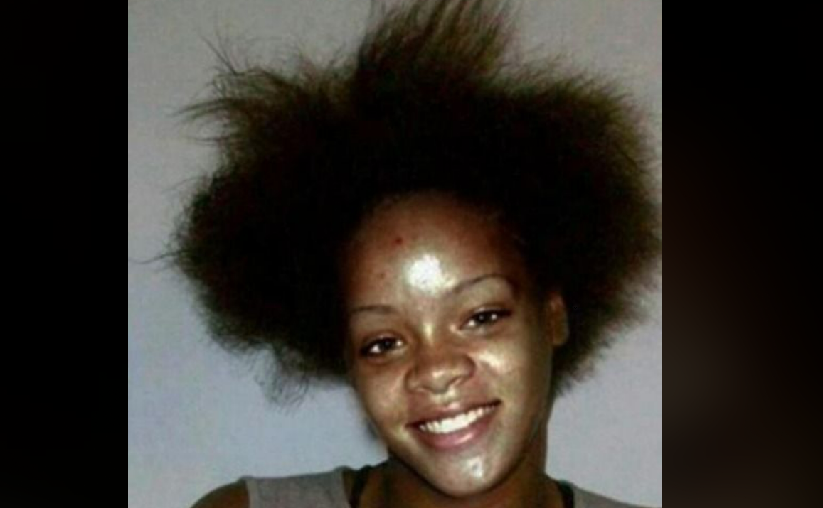 A woman with a wide smile and natural, voluminous hair stands against a plain background. Her skin appears shiny, and she is wearing a sleeveless top.